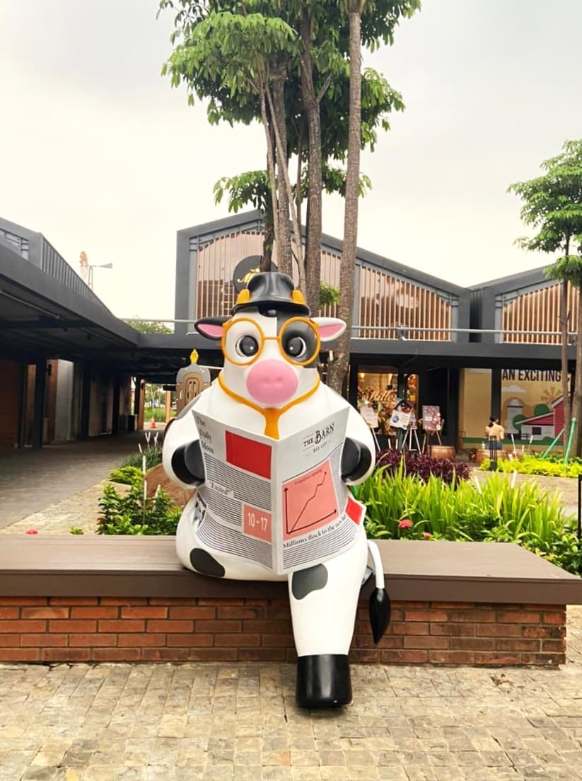 印尼 BSD City 的 The Barn 内,一尊戴眼镜和礼帽、坐在长椅上读报的大型奶牛雕像